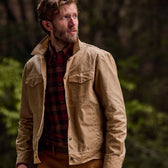 Bearded man wears Tom Beckbes Chatom Jacket over a plaid shirt, standing against a blurred natural backdrop.