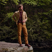 In a Chatom Jacket by Tom Beckbe and brown pants, a person stands on a rock ledge in the woods, looking out.