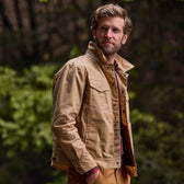 A bearded man in a Tom Beckbe Chatom Jacket stands outdoors surrounded by lush greenery.