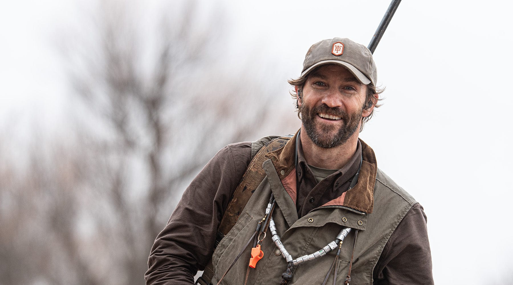 A man in outdoor gear and cap smiles, with a shotgun over his shoulder and a whistle lanyard around his neck.