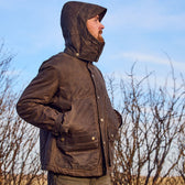 Someone wearing a hooded Fowler Jacket by Tom Beckbe stands outside with hands in pockets, against leafless shrubs.