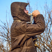 Person in a Tom Beckbe Fowler Jacket outdoors, using a duck call. Bare trees and clear blue sky behind.