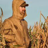 A person in a Tom Beckbe Fowler Jacket and cap stands in tall grass, gazing into the distance.
