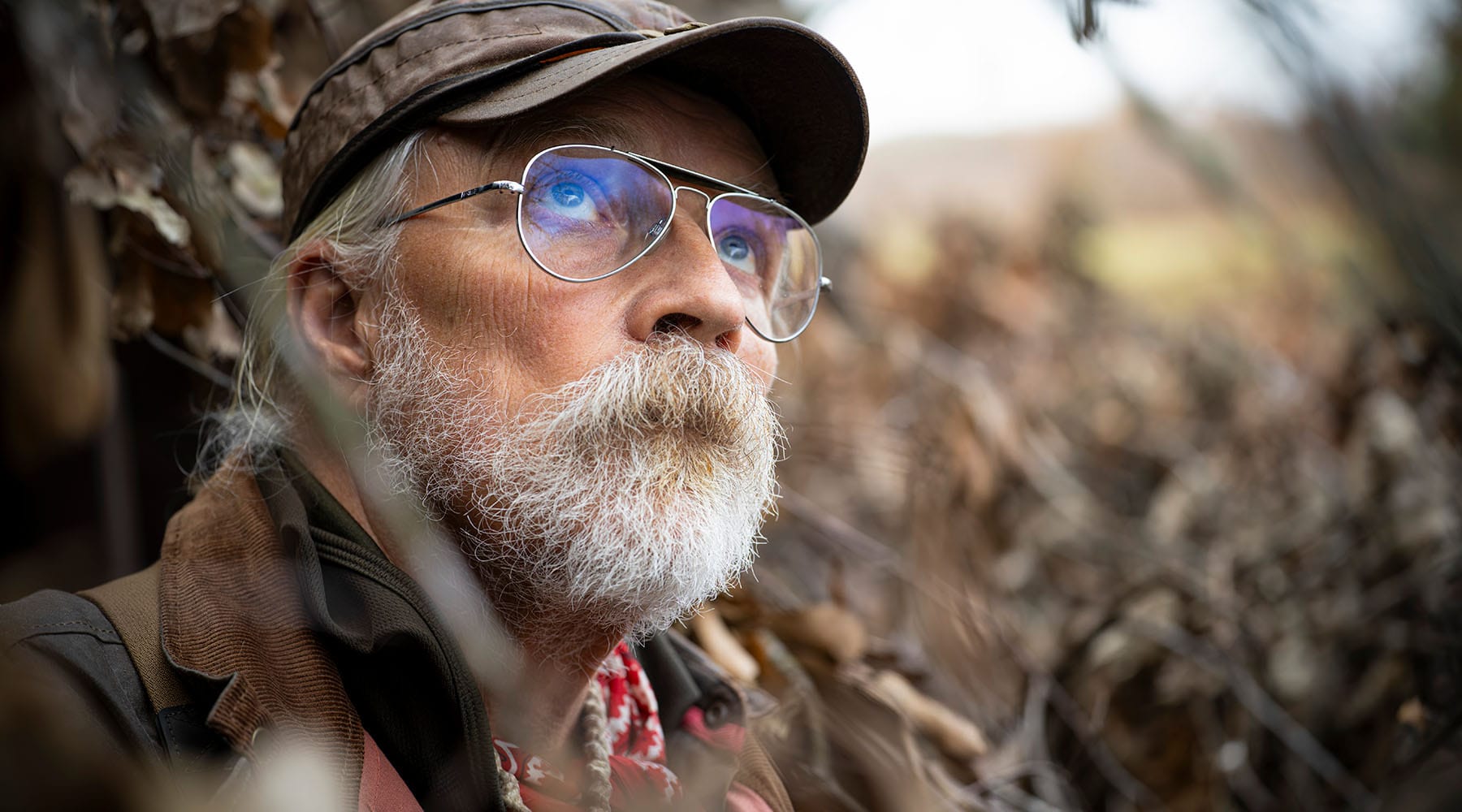 Older man with white beard, glasses, and cap, looking upward, surrounded by dried foliage outdoors.