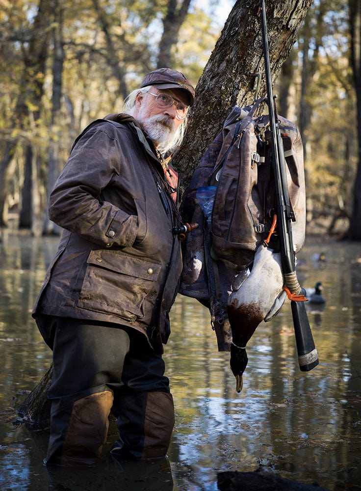 Older man in outdoor gear stands in shallow water near a tree, with a duck, backpack, and shotgun hanging from the tree.