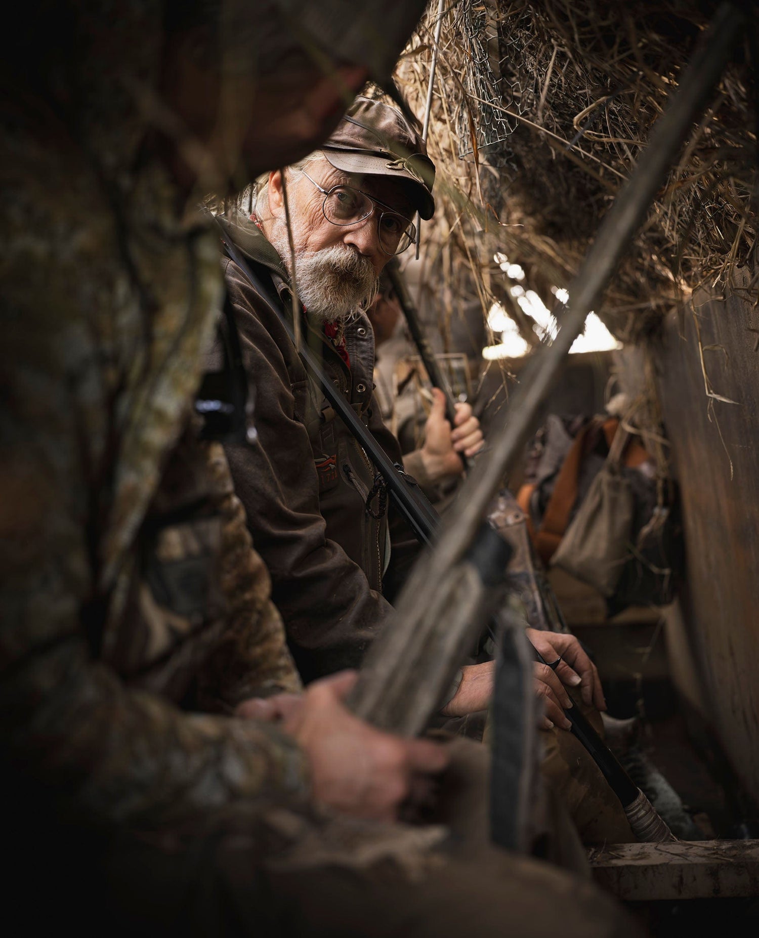 Older man in camouflage holds a shotgun while sitting in a hunting blind with other hunters.