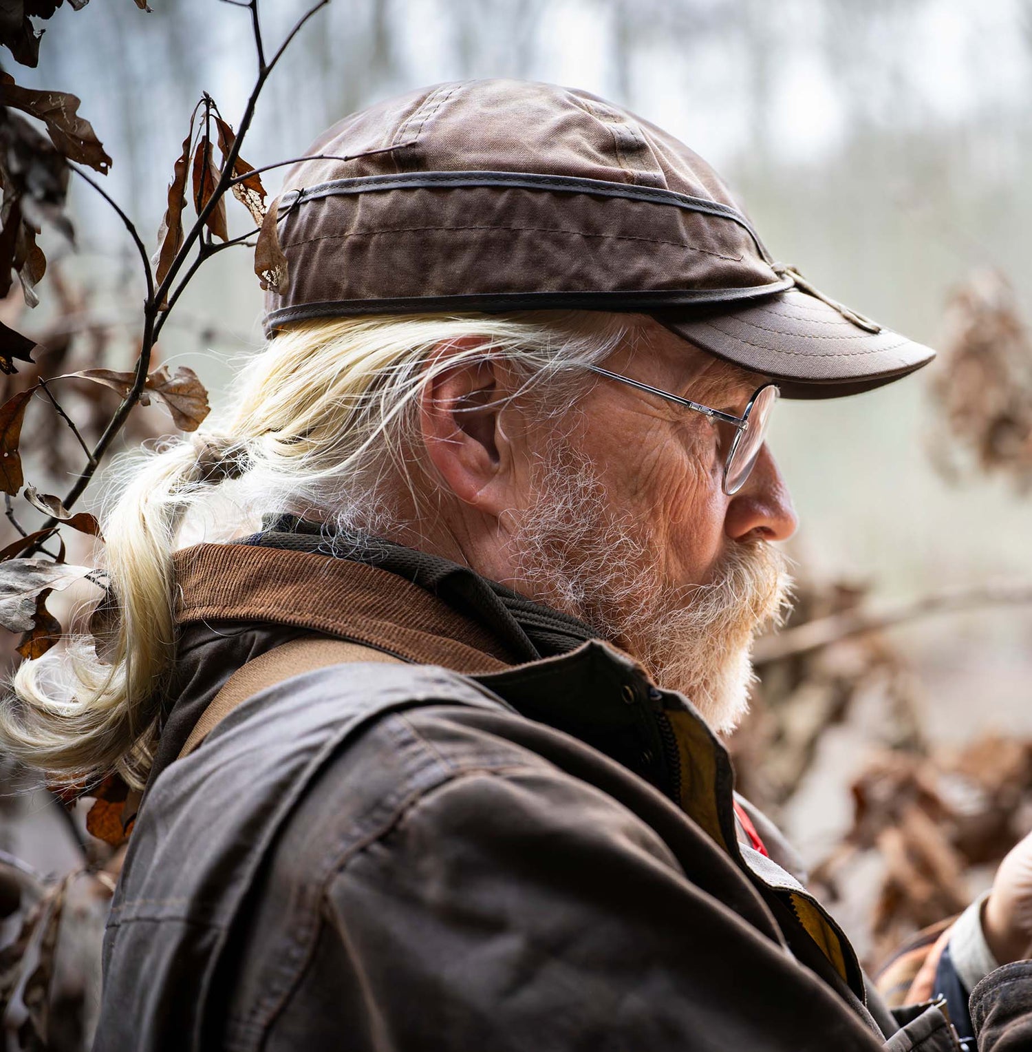 An older man with a gray beard and ponytail wears a brown cap and jacket while looking down outdoors.