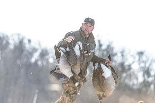 A man in outdoor gear holds two large geese by their necks with a blurred, wintry background.