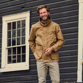 Man in tan Tom Beckbe Tensaw Jacket and khakis smiles in front of a dark wooden building with a window.