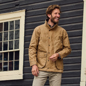 A man in a Tom Beckbe Tensaw Jacket and beige pants smiles in front of a dark wooden wall with a window.