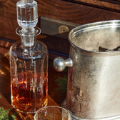 A Tom Beckbe Terrane No. 12 Decanter filled with amber liquid sits beside a silver ice bucket on wood in the sun.