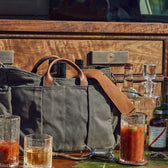 A Tom Beckbe Terrane No. 12 Decanter and jigger sit on a wooden surface, surrounded by handcrafted glassware and a canvas bag.