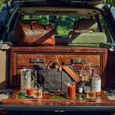 A car trunk holds a wooden bar setup with Tom Beckbe whiskey decanters, bottles, Terrane Glass, and bags in the sun.