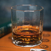 A glass of amber whiskey in a Tom Beckbe Terrane No. 12 Glass, on embossed leather with a book peeking from the background.