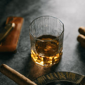 The Terrane No. 12 Glass by Tom Beckbe sits on a table with whiskey, a cigar, a brown wallet, and decor nearby.