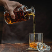 A hand pours amber liquid from a decanter into Terrane No. 12 Glasses by Tom Beckbe on wood, stopper nearby.