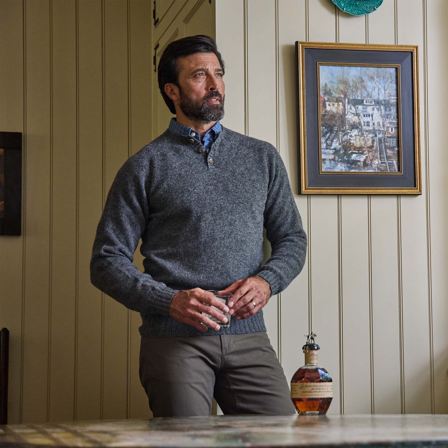 A man in a Tom Beckbe Albemarle Henley Sweater stands indoors near a table, with art on a cream-paneled wall behind him.
