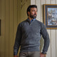 A man with dark hair and beard wears the Tom Beckbe Albemarle Henley Sweater, standing indoors by a framed painting.