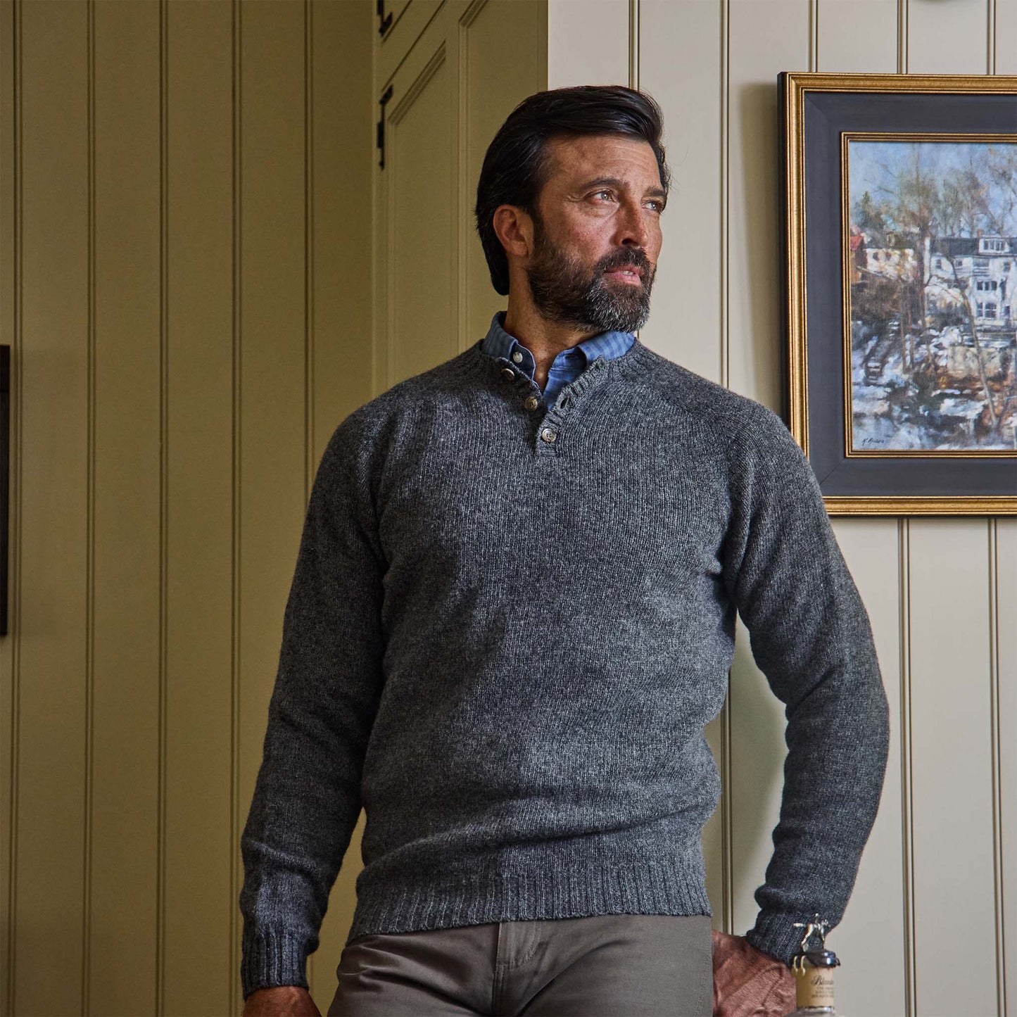 A man with dark hair and beard wears the Tom Beckbe Albemarle Henley Sweater, standing indoors by a framed painting.