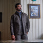A man wears the Tom Beckbe Albemarle Henley Sweater, standing indoors by wood paneling and a framed painting.