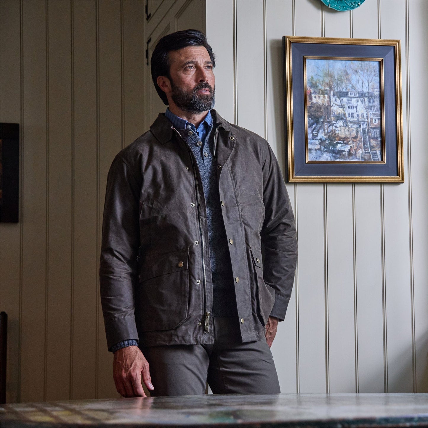A man wears the Tom Beckbe Albemarle Henley Sweater, standing indoors by wood paneling and a framed painting.