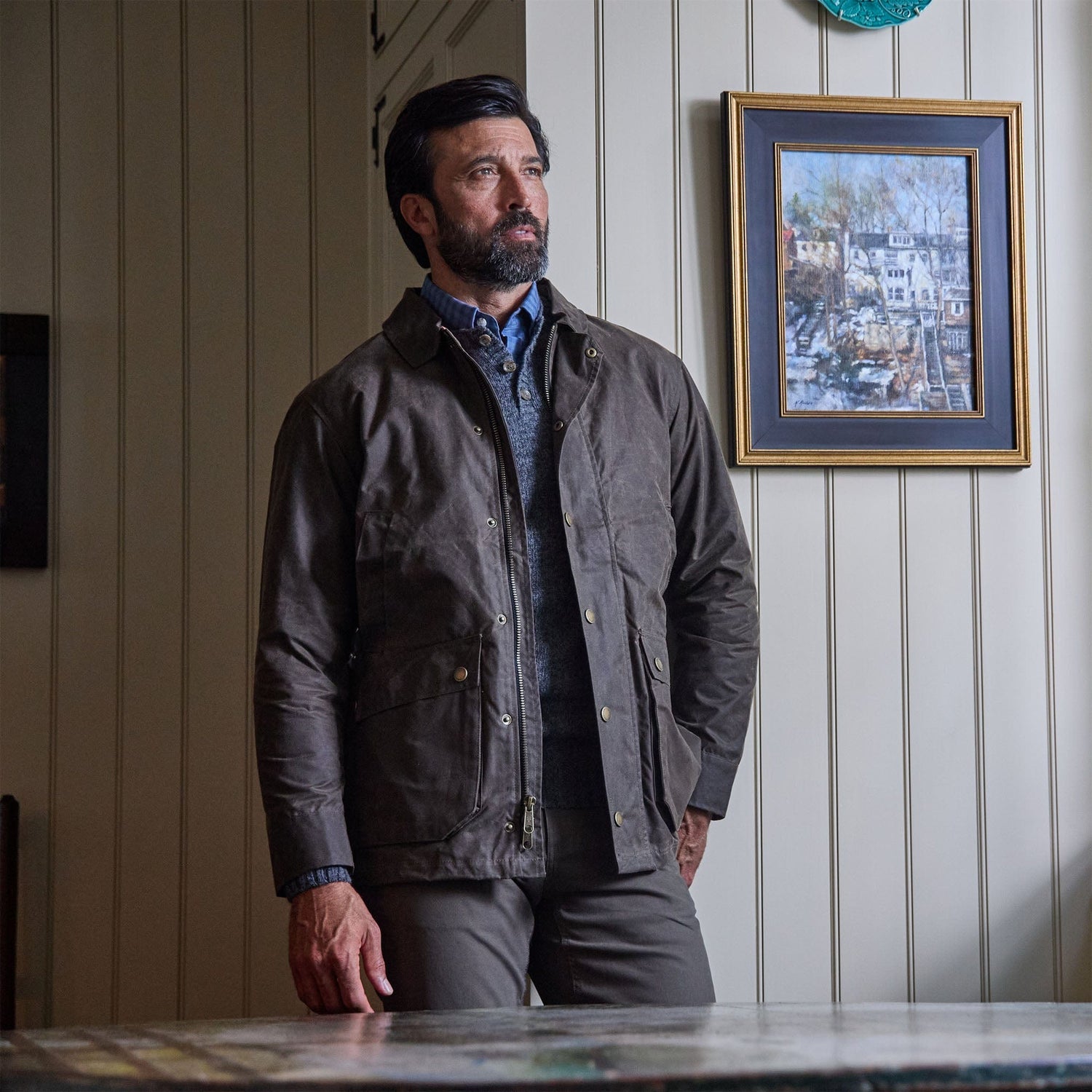 A man wears the Tom Beckbe Albemarle Henley Sweater, standing indoors by wood paneling and a framed painting.