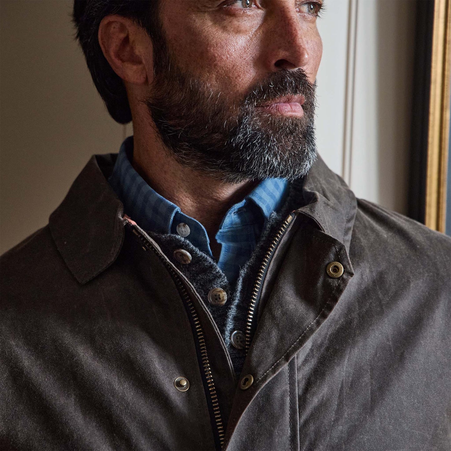 A bearded man wears the Tom Beckbe Albemarle Henley Sweater in grey over a blue shirt and brown jacket, looking sideways.