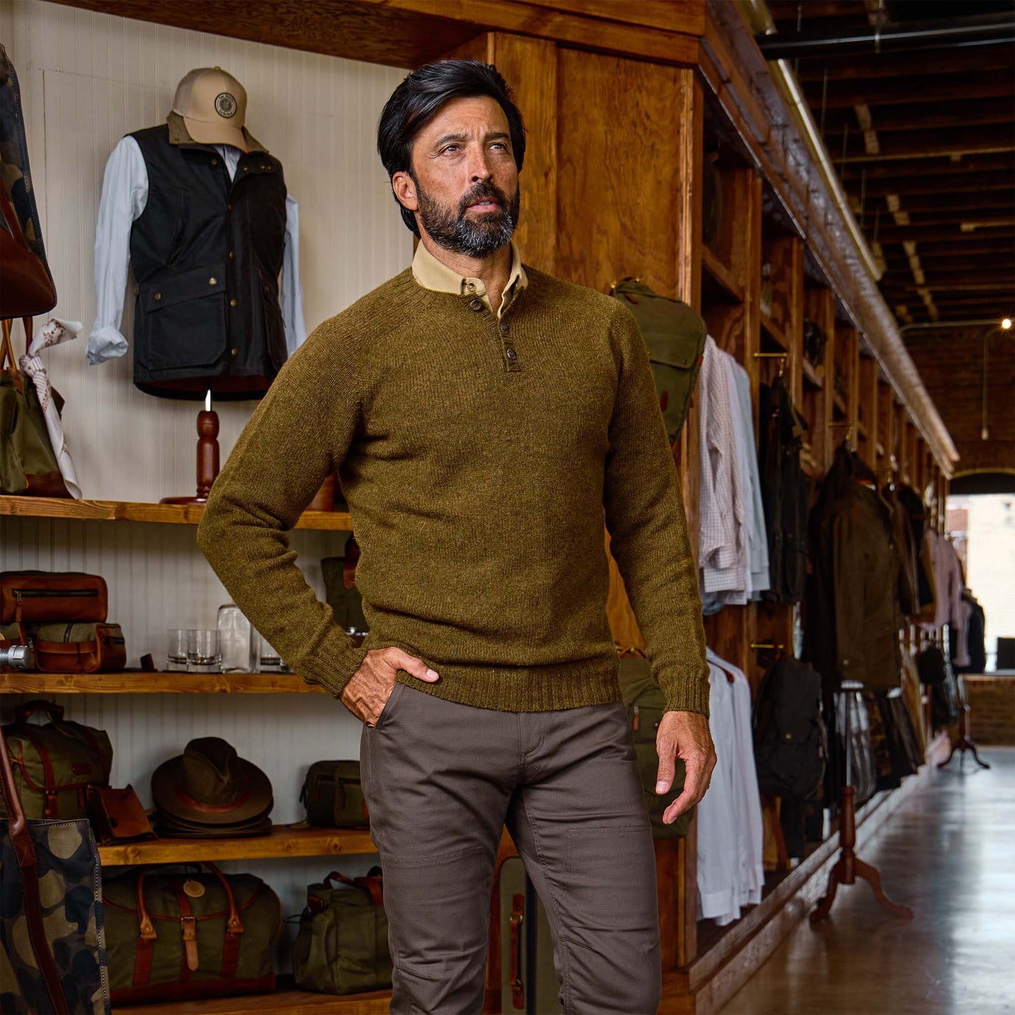 Man with dark hair and beard wears the Tom Beckbe Albemarle Henley Sweater in a store with wooden shelves.