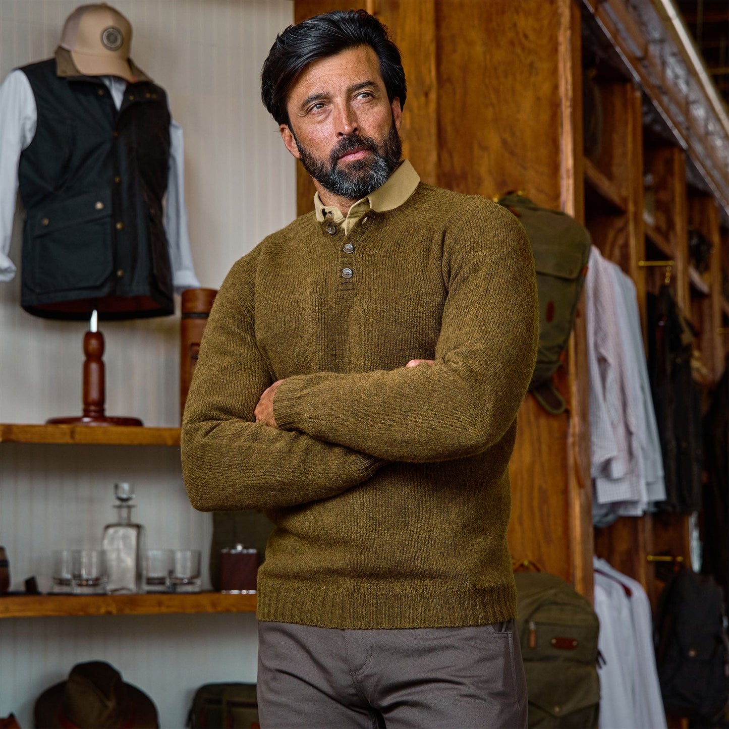 Man with dark hair and beard wears the Tom Beckbe Albemarle Henley Sweater in a store with shelves behind him.