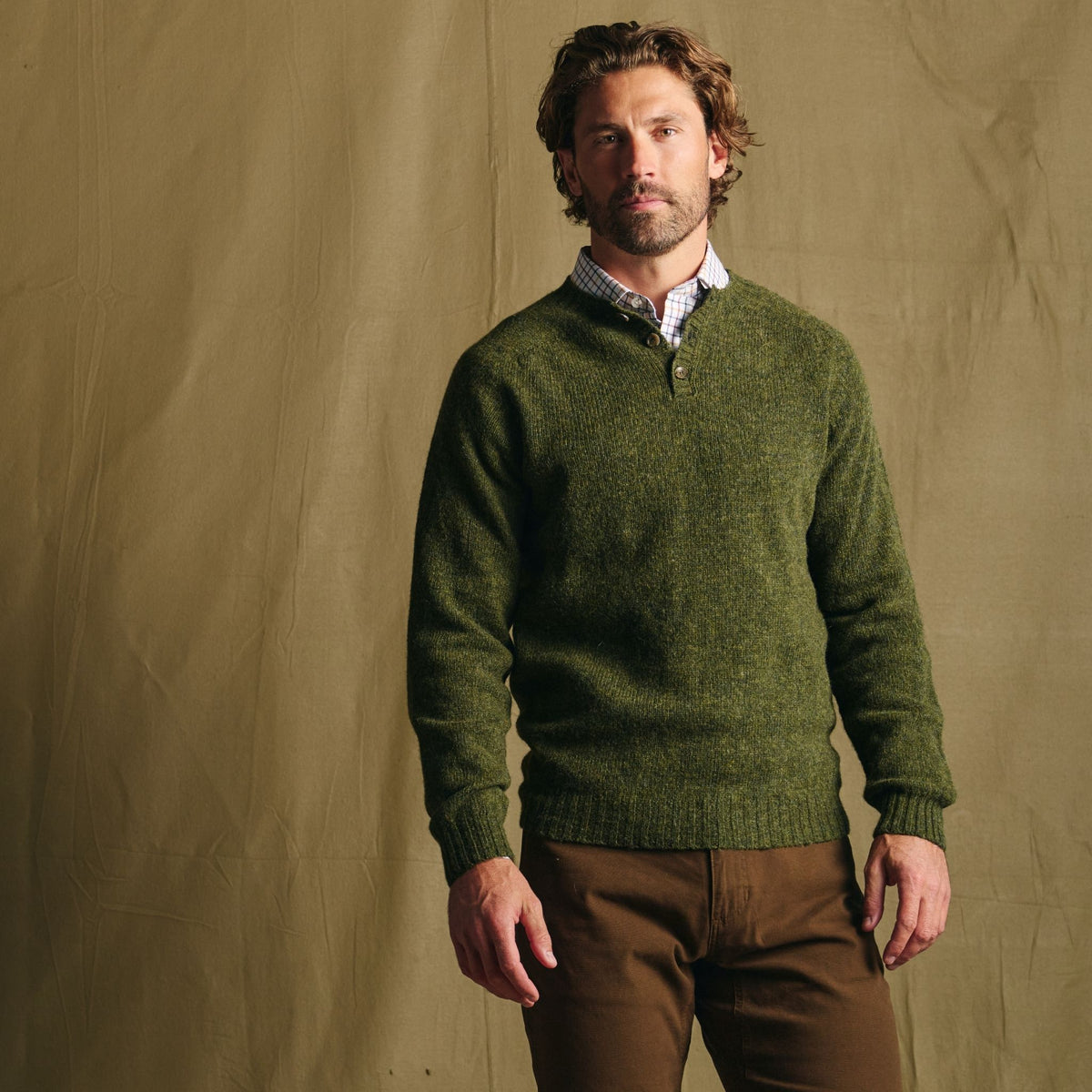 Man wears a Tom Beckbe Albemarle Henley Sweater over a collared shirt and brown pants against a beige backdrop.