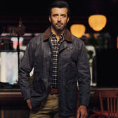Man in a Tom Beckbe Baldwin Jacket over plaid shirt stands indoors with warm lighting and blurred background.