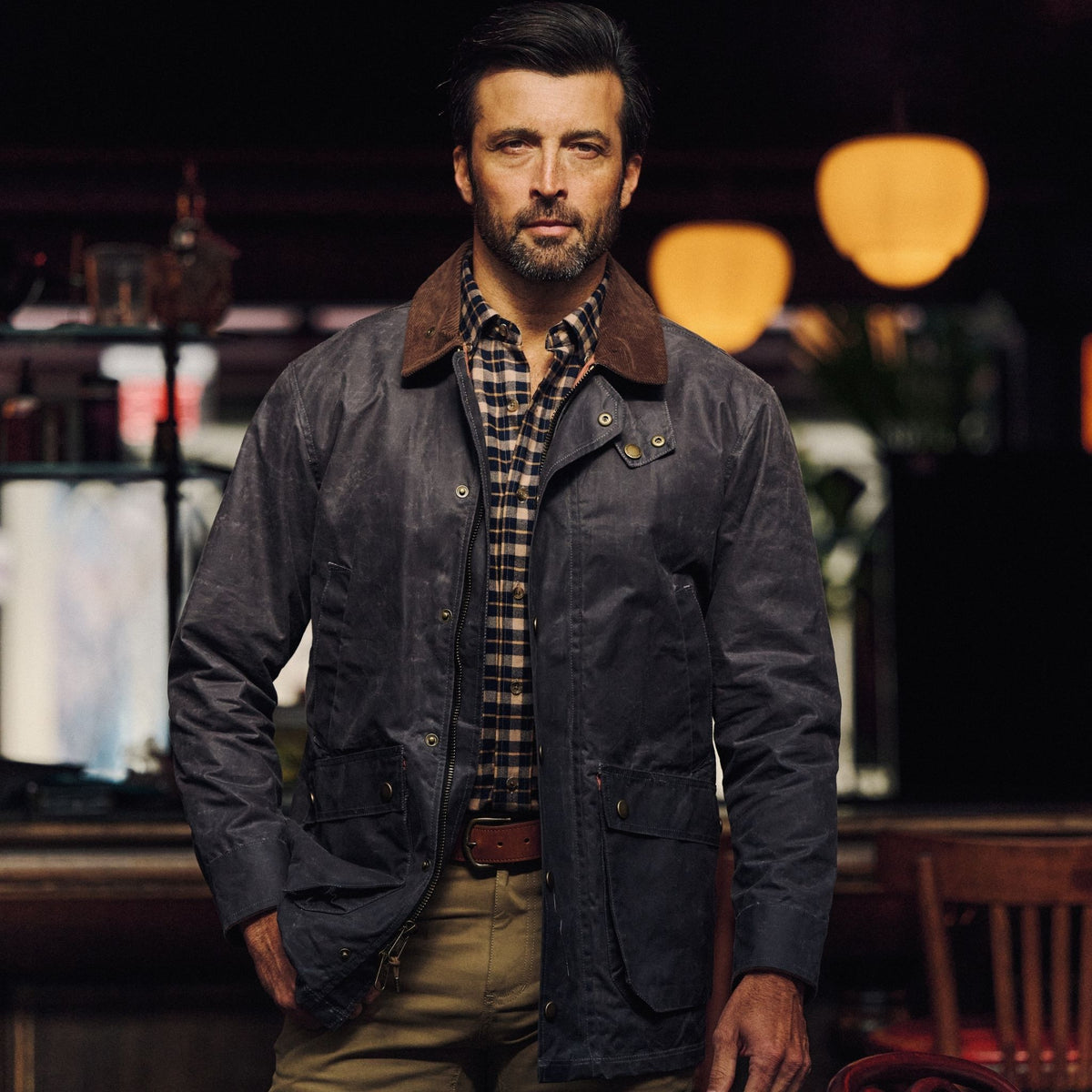 Man in a Tom Beckbe Baldwin Jacket over plaid shirt stands indoors with warm lighting and blurred background.