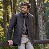 A man in a Tom Beckbe Braddock Quilted Jacket leans on a wooden fence in the forest.