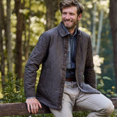 A man in a Tom Beckbe Braddock Quilted Jacket sits on a wooden fence in the forest, smiling.