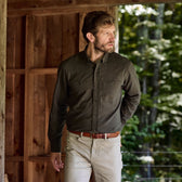 Man wears a Tom Beckbe Dixon Brushed Cotton Twill Shirt and light pants near a wooden structure with trees behind.