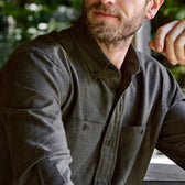 A bearded man in a Tom Beckbe Dixon Brushed Cotton Twill Shirt sits at a wooden table outdoors.