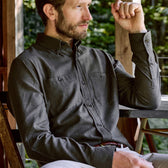 A man in a Tom Beckbe Dixon Brushed Cotton Twill Shirt sits outdoors, resting his arm on the table edge.