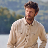 A bearded man with curly hair wears the Tom Beckbe Dixon Brushed Cotton Twill Shirt outdoors by trees and water.