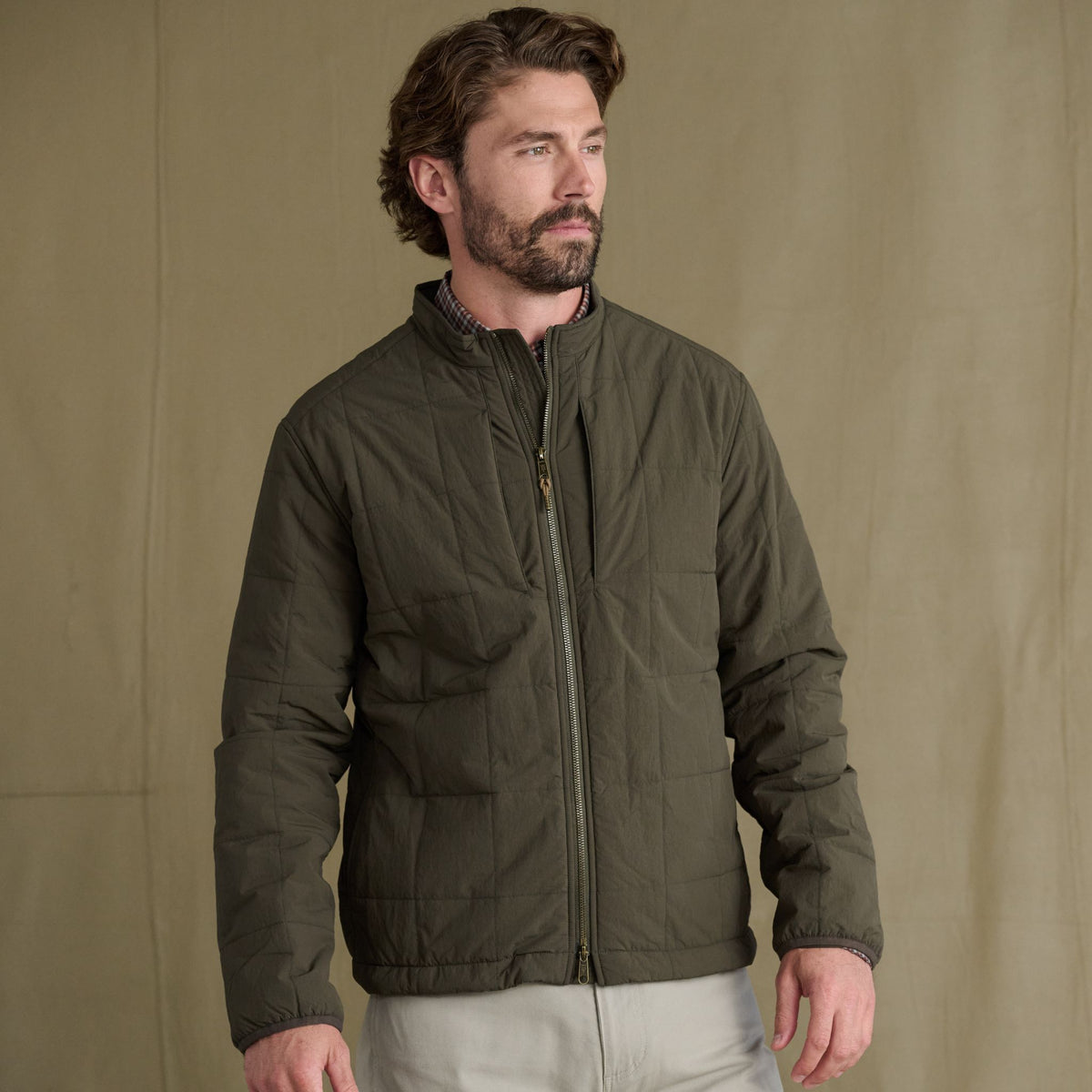 Man wears Tom Beckbe Burgess Quilted Jacket and light pants, standing against a plain tan background.