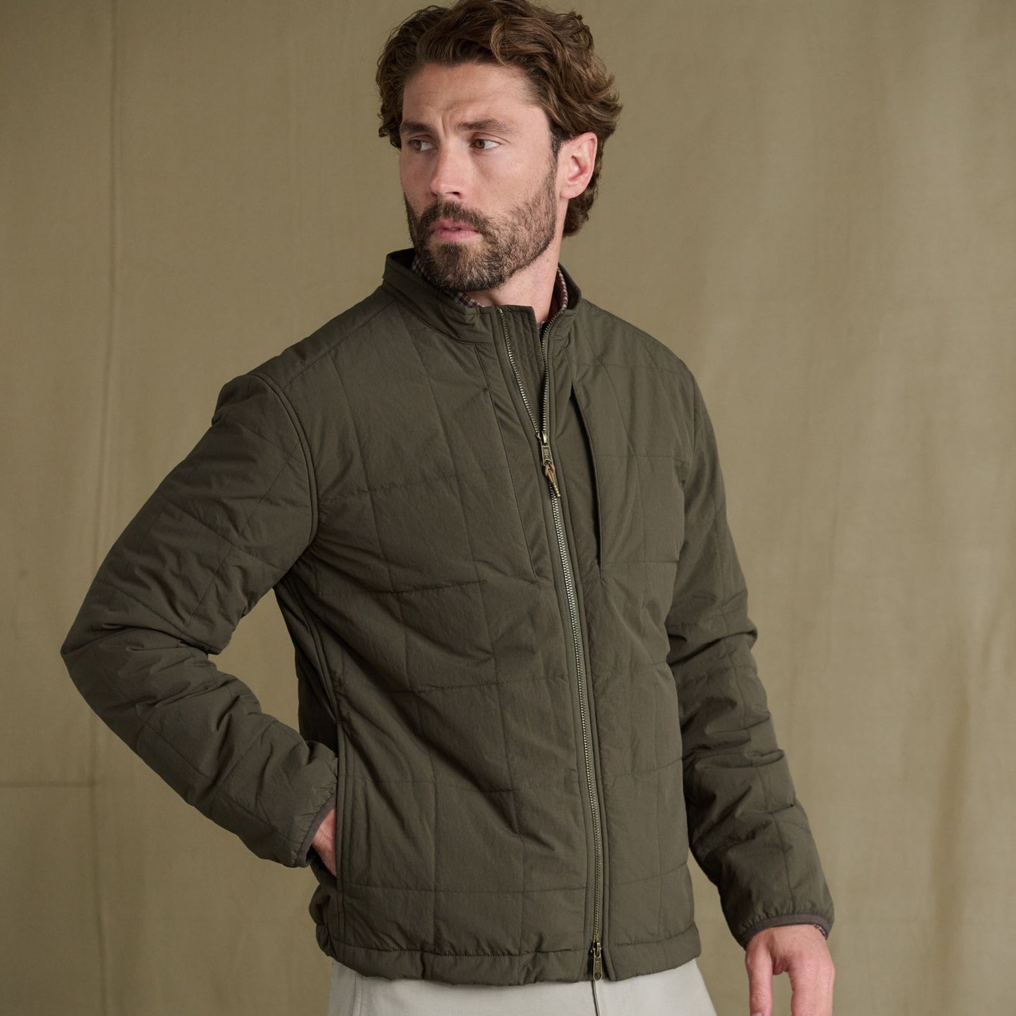 Man in Tom Beckbe Burgess Quilted Jacket with zip closure, standing against beige background, looking to the side.