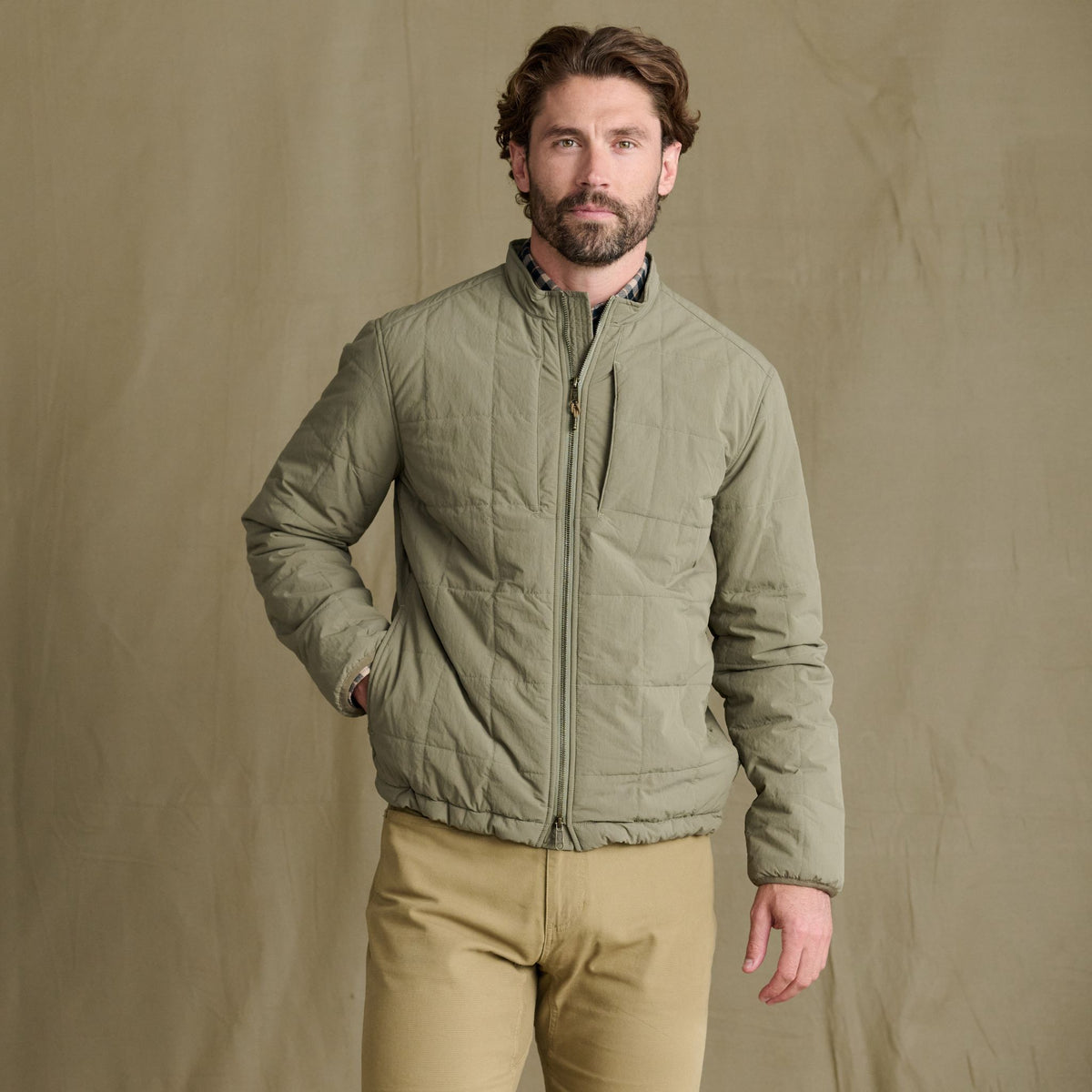 A bearded man wears the Tom Beckbe Burgess Quilted Jacket with khaki pants, standing before a beige background.