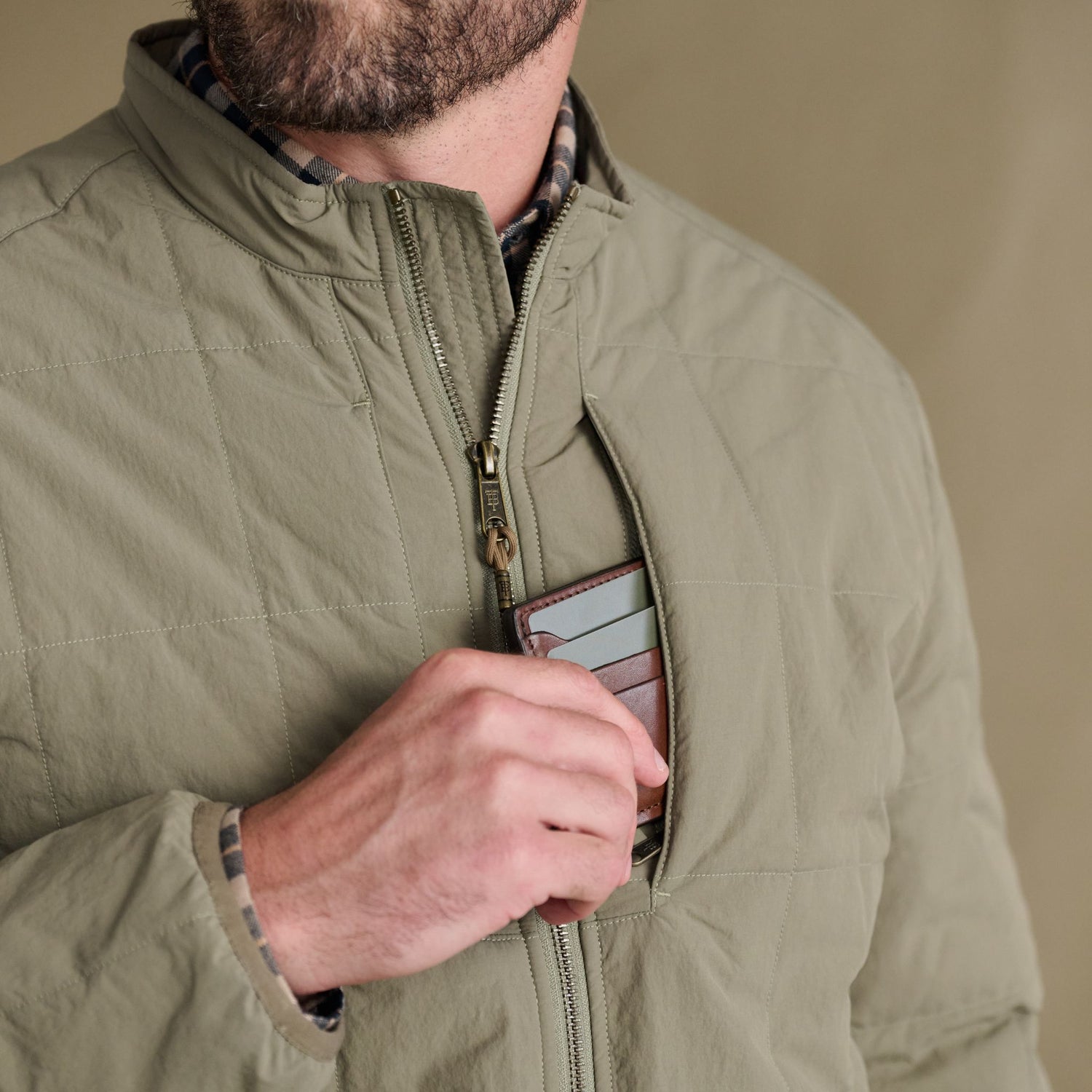Man slips a wallet into the chest pocket of Tom Beckbe’s Burgess Quilted Jacket in light olive for secure style.