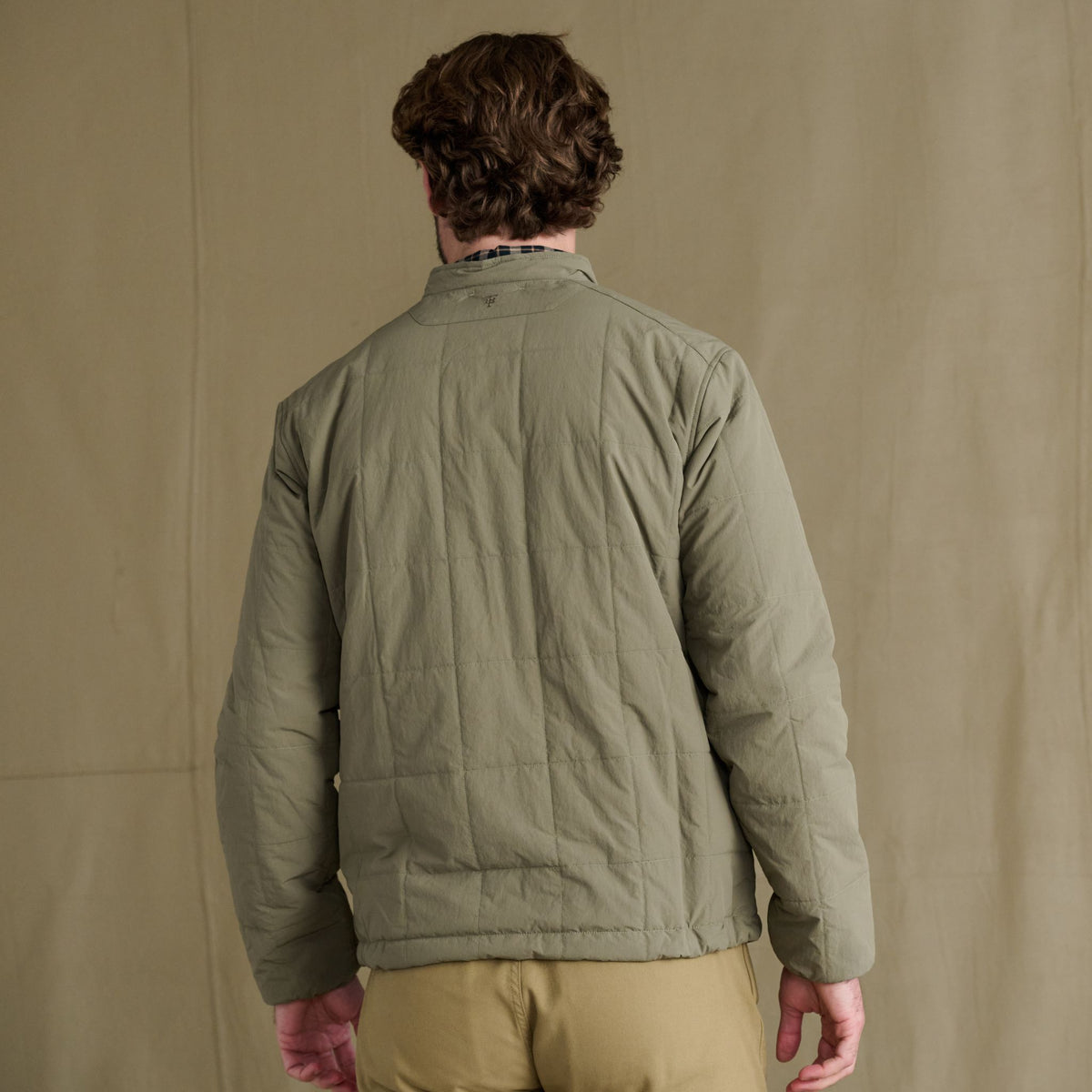 A person faces away, wearing the Tom Beckbe Burgess Quilted Jacket with khaki pants against a beige background.