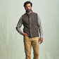 Man in a Tom Beckbe Burgess Quilted Vest over a patterned shirt and tan pants, against a light green background.