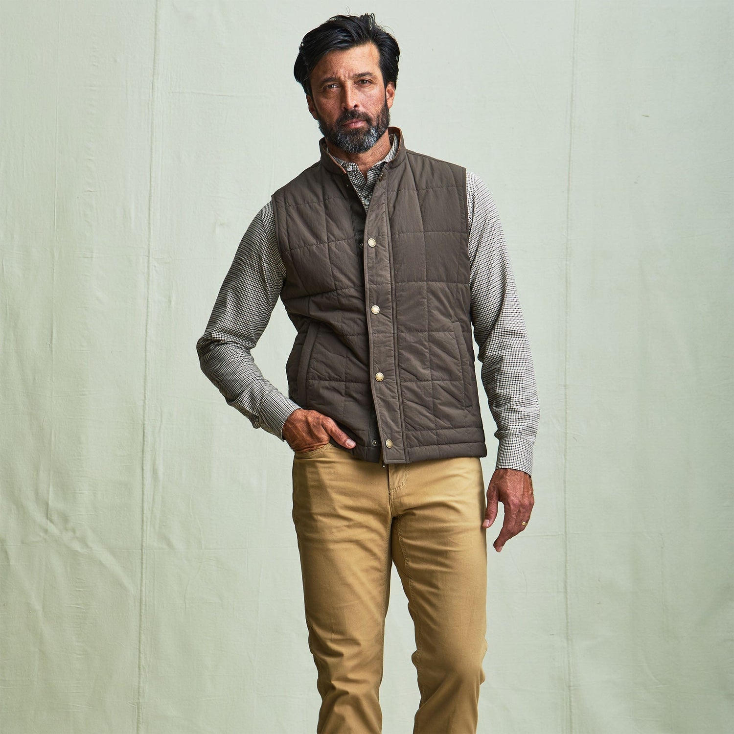 Man in a Tom Beckbe Burgess Quilted Vest over a patterned shirt and tan pants, against a light green background.