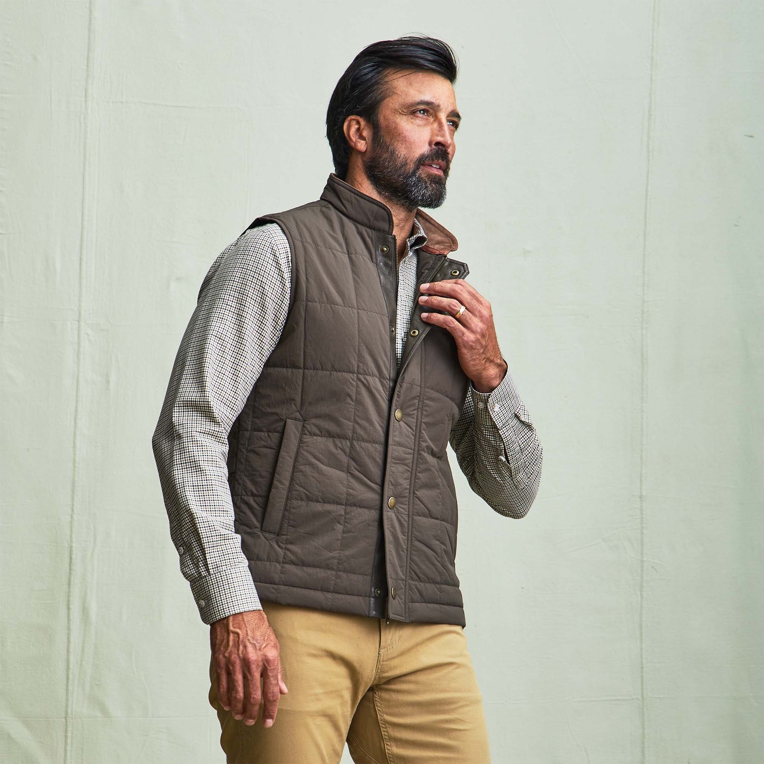 Man wears the Tom Beckbe Burgess Quilted Vest over a checkered shirt and khaki pants against a plain light background.