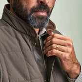 A man in a Tom Beckbe Burgess Quilted Vest and checked shirt adjusts his collar, revealing a gold wedding ring.