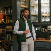 Man in Tom Beckbe Burgess Quilted Vest holds a glass by a glassware cabinet, surrounded by bookshelves and art.