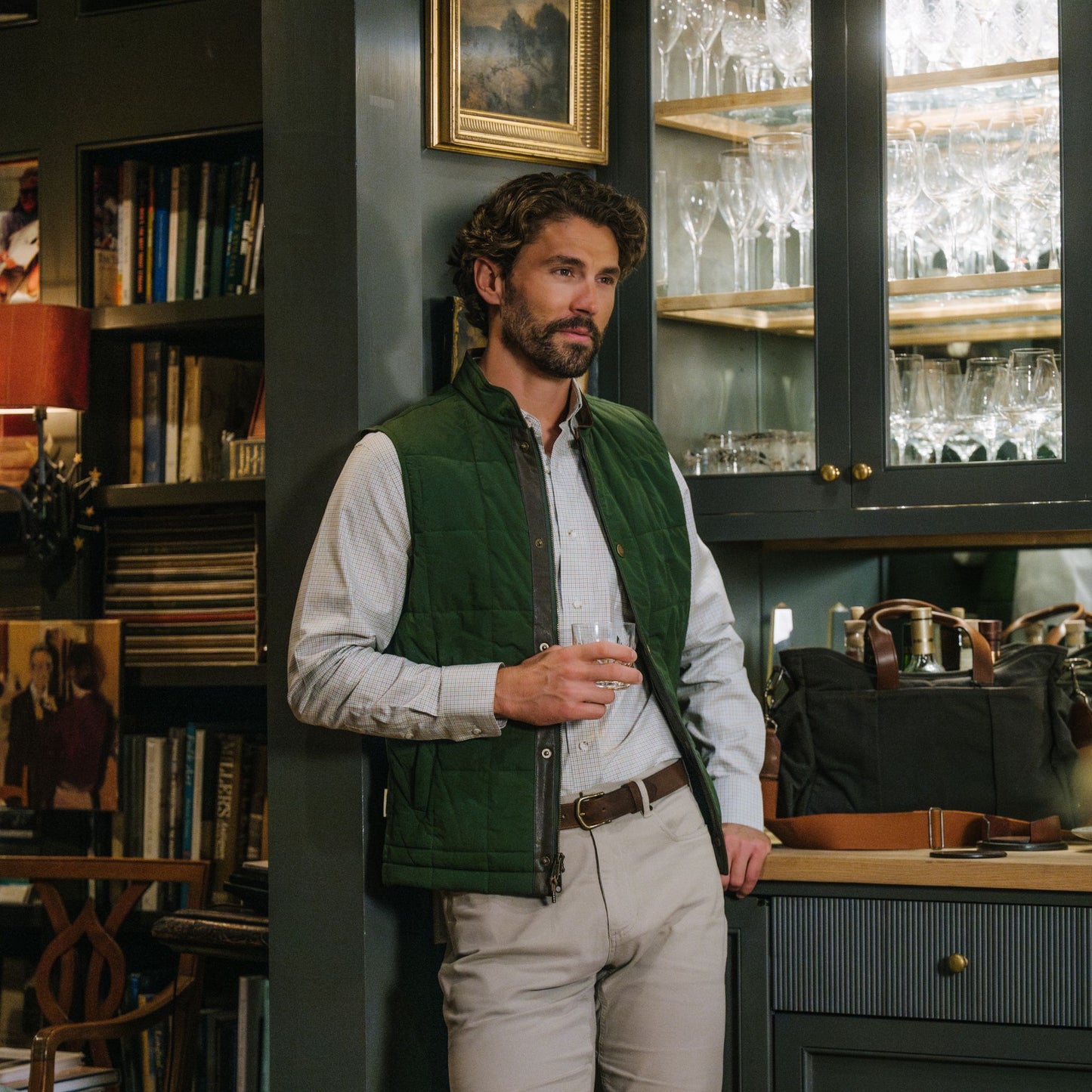 Man in Tom Beckbe Burgess Quilted Vest holds a glass by a glassware cabinet, surrounded by bookshelves and art.