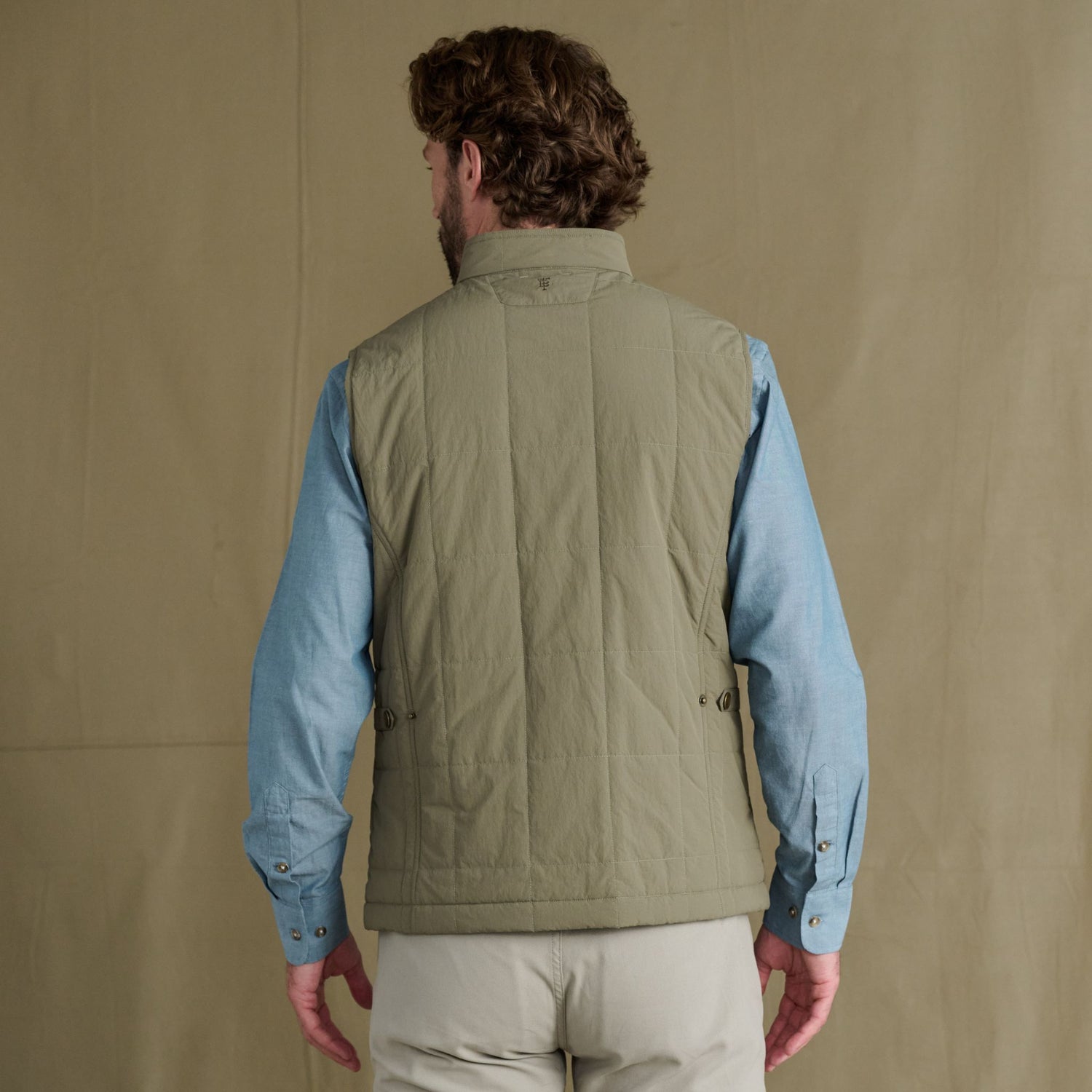 Man in Tom Beckbe Burgess Quilted Vest over blue shirt, beige pants, stands against neutral background, facing away.